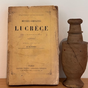 May include: An antique book titled "OEUVRES COMPLÈTES DE LUCRÈCE" with French translation, next to a brown, handcrafted ceramic vase. The book has a worn, aged appearance with visible text. The vase is cylindrical with a flared top.