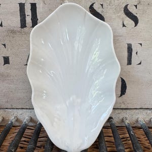 May include: A white, shell-shaped ceramic serving dish. The dish has a scalloped edge and a glossy finish. The background features a weathered wooden surface with black stenciled letters and a woven texture.