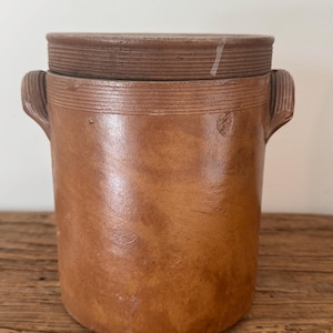 May include: A cylindrical brown stoneware crock featuring a matching lid, two side handles, and horizontal ribbed detailing around the upper rim. The vessel has a smooth, mottled finish with a small circular indentation on the front surface.