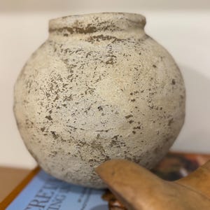 May include: A round, off-white ceramic vase with a textured surface and a slightly darker rim. The vase has a bulbous shape and a small opening at the top. The vase is a decorative item for home decor.