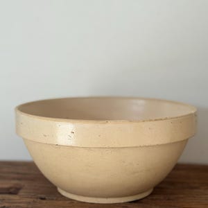 May include: A round, cream-colored ceramic mixing bowl with a wide, flat rim and a smooth, glazed finish. The bowl features a deep, conical shape with a small, circular base, showcasing a simple, functional design with subtle surface texture.