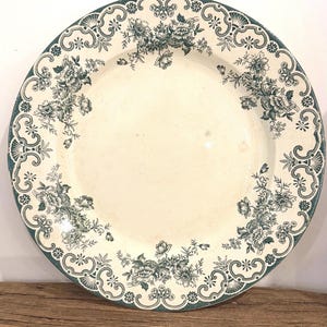May include: A round ceramic plate with an off-white center and a wide border featuring a dark green floral pattern. The rim is decorated with an intricate scrollwork design that frames clusters of detailed flowers and leaves.