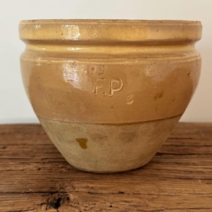 May include: A tan-colored ceramic crock with a glossy upper glaze and a matte lower section. The vessel features a wide, rounded rim and a distinct horizontal groove below the rim. The letters F.P are embossed on the side of the exterior surface.