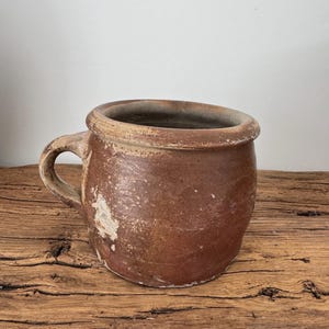 May include: A rustic, brown ceramic mug with a handle. The mug has a wide, rounded body and a slightly flared rim. The exterior shows signs of wear, with a mottled texture and areas of lighter color. The mug sits on a wooden surface.