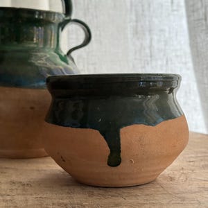 May include: A small, handcrafted terracotta pot with a dark green glazed rim and a drip design. The pot has a rounded shape and sits on a wooden surface. A larger green glazed pitcher is partially visible in the background.