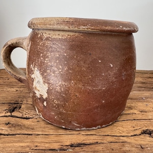 May include: A rustic, brown ceramic crock with a lid and handle. The crock has a rounded shape and a textured surface with areas of lighter color. The handle is a simple loop, and the lid sits flush on top. The piece is likely vintage.