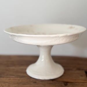 May include: A white ceramic pedestal cake stand featuring a shallow, wide bowl with a scalloped edge and a raised, embossed leaf and vine pattern around the exterior rim. The stand has a smooth, flared base and a central column support.
