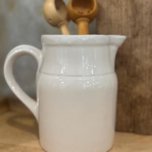 May include: A small, off-white ceramic pitcher with a handle and spout. Two wooden scoops are placed inside the pitcher. The background includes a wooden surface and a blurred background.