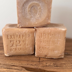 May include: Three stacked light brown square soap blocks. The top block features a horseshoe emblem. The bottom left block reads EXTRA PUR 72% D'HUILE. The bottom right block reads J.B. PAUL MARSEILLE 500 GRAMMES. The soap has a matte, textured finish.
