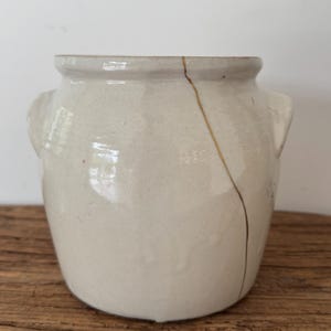 May include: A cream-colored ceramic jar with two small handles and a wide opening. A gold-colored line runs down the side, indicating a repair. The jar sits on a wooden surface.