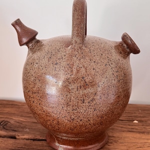 May include: A speckled brown ceramic pitcher with a rounded body, two spouts, and a handle. The pitcher has a wide base and a textured surface, giving it a rustic appearance. The spouts are angled and the handle is arched.