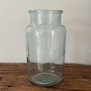 Op de afbeelding: Een heldere glazen pot met een brede cilindrische body en een iets smallere hals. De pot is transparant, waardoor de inhoud zichtbaar is. Hij staat op een houten oppervlak, met een effen muur op de achtergrond.