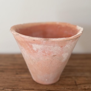 May include: A conical terracotta plant pot with a wide, open rim and a weathered, matte finish. The surface displays a natural, earthy orange-brown color with patches of white mineral deposits and a slightly uneven, textured clay construction.