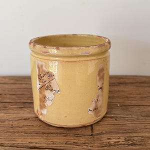 May include: A yellow ceramic jar with a slightly distressed appearance. The jar has a rounded shape with a wide opening and a narrow rim. Faded text is visible on the jar's surface, adding to its vintage aesthetic. The jar is placed on a wooden surface.