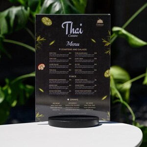 May include: A black Thai Cuisine menu with white text, featuring starters, salads, rice, and drink options. The menu is displayed on a white table with a black stand, with green foliage in the background.