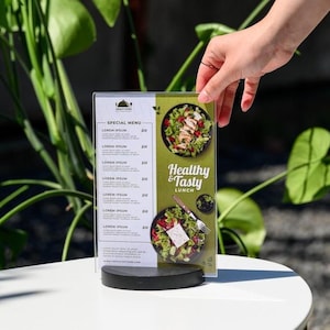 May include: A restaurant menu with a special menu section and a "Healthy & Tasty Lunch" section. The menu features images of salads. The menu is held by a hand and is on a white table with green plants in the background.