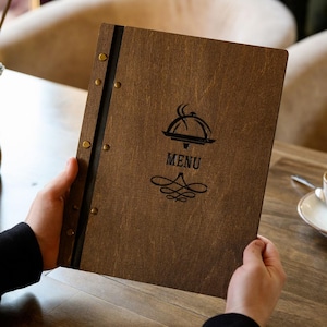 May include: A wooden menu with the word "MENU" printed in black, along with a food service graphic. The menu is held open, revealing the interior pages. The cover is a dark brown color, with brass rivets along the spine.