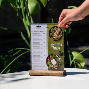 May include: A clear acrylic menu with a wooden base, featuring a "Special Menu" section and a "Healthy & Tasty Lunch" advertisement with images of salads. The menu is held by a hand.