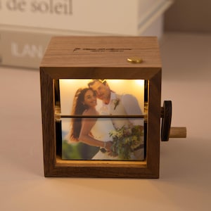 May include: Wooden photo display box with a warm light illuminating a wedding photo. The box has a crank on the side and a gold-colored button on top. The photo shows a couple in wedding attire. The box is made of wood.