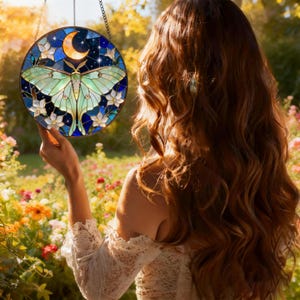 May include: A round stained glass suncatcher featuring a luna moth design. The moth has green wings and is set against a blue background with a crescent moon and stars. White flowers surround the moth. The suncatcher hangs from a chain.