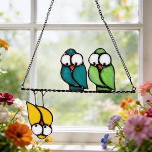 Puede incluir: Atrapasoles de pájaros de vidrieras con tres pájaros. Dos pájaros están posados en una barra de metal negro, uno turquesa y otro verde. Un pájaro amarillo cuelga debajo. Los pájaros tienen ojos blancos grandes y picos naranjas.