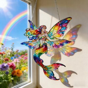 Può includere: Un acchiappasole a forma di fata sirena in vetro colorato, con grandi ali dettagliate. La fata ha capelli arcobaleno e una coda abbinata. Appeso a una catena d'argento, proietta un'ombra arcobaleno. Sullo sfondo si vedono un arcobaleno e dei fiori.