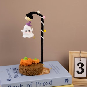 May include: A handmade Halloween-themed crochet lamp featuring a white ghost with a purple hat, two orange pumpkins, and a black lamp post. The lamp is decorated with small colorful beads and sits on a brown crocheted base. The lamp is approximately 10 inches tall.