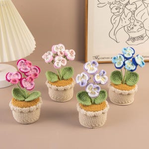 May include: Four crocheted flower pots, each with a different color of flowers. The flowers are pink, light pink, purple, and blue. The pots are beige with a yellow-brown base and green leaves.