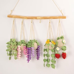 May include: A collection of six crochet hanging planters with various plant designs. The planters are suspended from a wooden rod with beige cords. The plants include green vines, pink and purple flowers, yellow sunflowers, white daisies, and red strawberries.