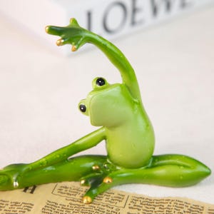Könnte beinhalten: Eine grüne Froschfigur in Yoga-Pose, mit erhobenen Armen und gespreizten Beinen. Der Frosch hat schwarze Augen und goldene Akzente an Händen und Füßen. Die Figur steht auf einer Oberfläche mit Zeitungspapier.