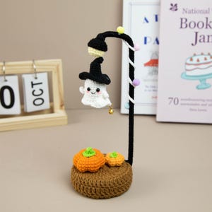 May include: A handcrafted Halloween-themed crochet decoration featuring a smiling ghost wearing a witch hat under a black lamp post. Two orange pumpkins sit on a brown crocheted base. The date on the calendar is October 10.