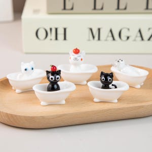 Puede incluir: Una colección de pequeñas bañeras de cerámica blanca, cada una con una figura de animal diferente. Las figuras incluyen gatos, un oso y una foca. Los gatos son negros y blancos, y el oso tiene una cereza roja en la cabeza.