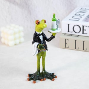May include: A whimsical figurine of a frog dressed as a waiter, wearing a black tuxedo and holding a tray with a green wine bottle and a glass. The frog has orange eyes and green skin. The figurine is a decorative item.