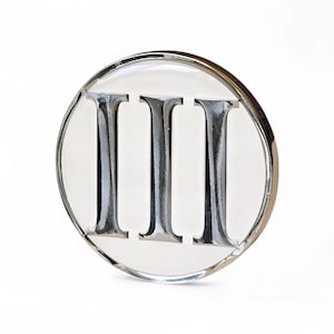 Peut inclure: Un objet clair et circulaire avec un bord métallique argenté. L'objet présente le chiffre romain « III » dans une police argentée et réfléchissante, centré sur un fond blanc. Le design est simple et élégant, avec une finition polie.