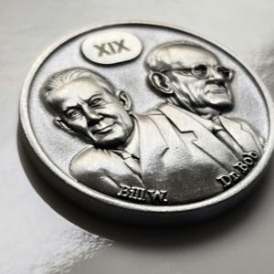 May include: A silver coin featuring bas-relief portraits of two men, one wearing glasses. The coin has the text "XIX", "Bill W.", and "Dr. Bob" inscribed. The coin has a detailed, textured surface.