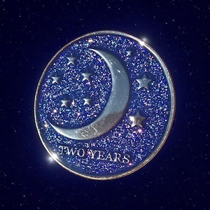 Könnte beinhalten: Runde, blaue und silberne Gedenkmünze mit Mond und Sternen. Der Hintergrund ist glitzernd, und am unteren Rand steht "TWO YEARS".
