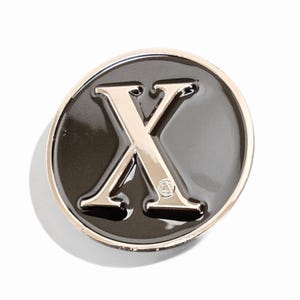 May include: A round, black enamel pin with a gold-colored border. The pin features a gold-colored letter "X" in the center. A small symbol is visible at the bottom of the letter. The pin is set against a white background.