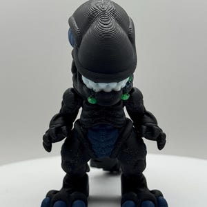 May include: A black 3D-printed dinosaur figurine with blue detailing on the eyes, belly, and toes. The figurine has a textured surface and a stylized design, with a wide head and small arms.