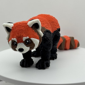 May include: A 3D-printed red panda figurine with a detailed textured surface. The figurine features a red body, black legs, and a striped tail. The panda has white accents on its face and ears, and brown markings around its eyes.