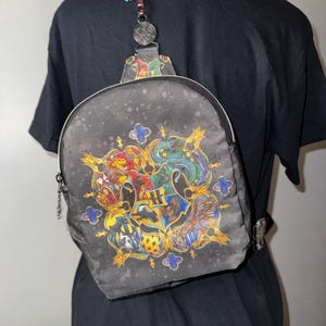 May include: A black mini backpack featuring a colorful, detailed crest design on the front. The crest includes four distinct sections with animal motifs and a central letter H. The bag has a single strap with a metallic attachment and a zipper closure.