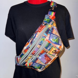Può includere: Un marsupio a tracolla con una stampa colorata di francobolli in stile vintage che raffigurano creature mitiche e luoghi come Bigfoot, Jackalope e Haleakala. La borsa ha due scomparti con cerniera grigia e una tracolla arcobaleno.