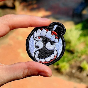 May include: Embroidered patch featuring a cartoon bomb with a smiling face, large eyes, and a black top hat. The patch has a white border and is held up against a blurred green background.