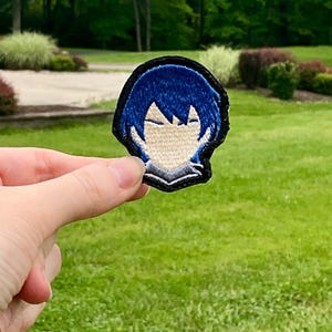 May include: Embroidered patch featuring a stylized anime character's face with blue hair and a black border. The patch is held against a backdrop of green grass and trees.