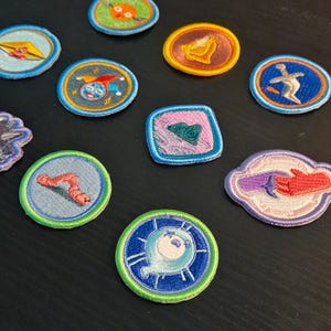 May include: A collection of ten embroidered fabric patches in various shapes and colors. The designs include a jester, a sword, a worm, a spiked mace, a heart, a bell, and other circular icons with vibrant thread work and stitched borders.