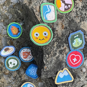 May include: A selection of embroidered patches featuring diverse designs such as a skull, a mountain, a snowflake, and a '5K' logo. The patches are colourful with stitched borders, in various shapes and sizes.