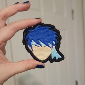 May include: An embroidered patch featuring a stylized anime character head with spiky dark blue hair, a light blue headband, and a tan face. The design is outlined with a thick black border, creating a distinct graphic shape for this textile accessory.