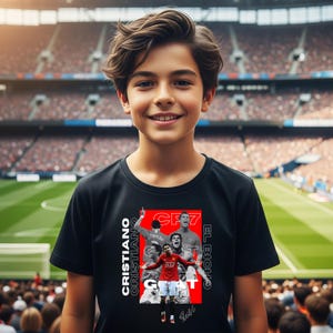 May include: A young person wearing a black t-shirt featuring a graphic of Cristiano Ronaldo in a red jersey. The shirt includes the text "CRISTIANO" and "EL BICH0" in white, with the number "7" and the word "GOAT". The background shows a stadium.