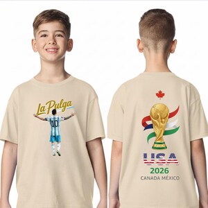 May include: Beige t-shirt featuring a soccer player graphic with the text "La Pulga" on the front. The back of the shirt displays the FIFA World Cup trophy, the USA flag, and the text "2026 CANADA M&Eacute;XICO".