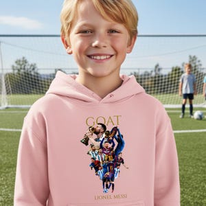 May include: A light pink hoodie featuring a collage of Lionel Messi images and the word "GOAT" in gold lettering. The hoodie has a front pocket and a hood. The text "Lionel Messi" is printed at the bottom of the graphic.