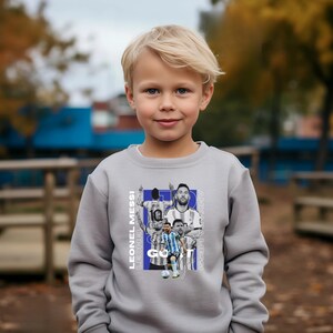 May include: A grey sweatshirt with a graphic of Lionel Messi. The design includes the text "LEONEL MESSI" and "GOAT" in white and blue. The sweatshirt is worn by a young child, with a blurred outdoor background.